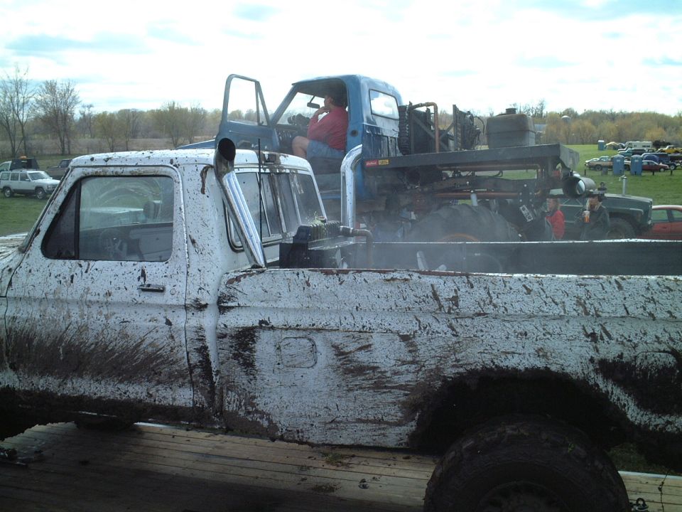Mud-caked white Ford pickup at a truck-pull event, driver in red shirt behind a blue cab