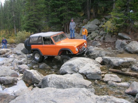 1995 Ford Bronco - An orange Ford Bronco traverses a challenging rocky stream, with onlookers observing the off-road adventure.