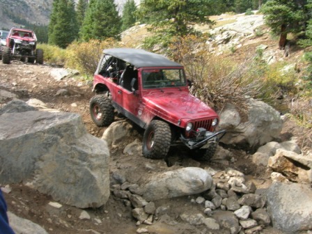 1995 Ford Bronco - Two off-road vehicles tackle a challenging, boulder-strewn path in a mountainous, wooded area.