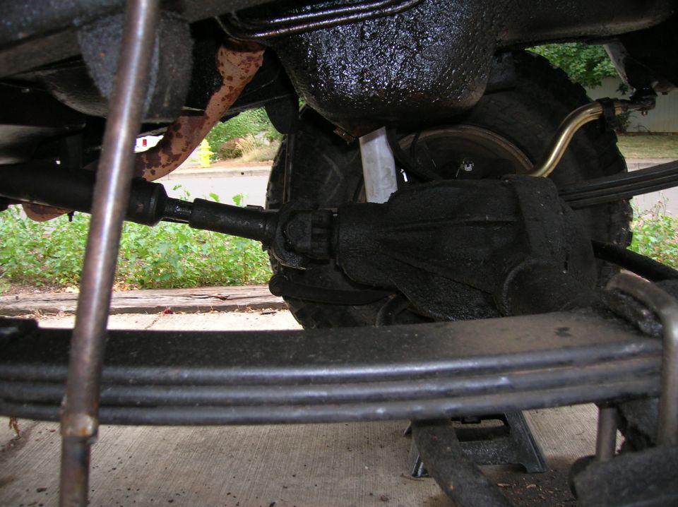 Underside view of a truck's rear axle, leaf springs, and driveshaft. A Supermotors logo is visible at the bottom right