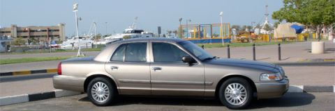 2007 Mercury Grand Marquis - A gold sedan is parked near a waterfront area with boats and buildings visible.