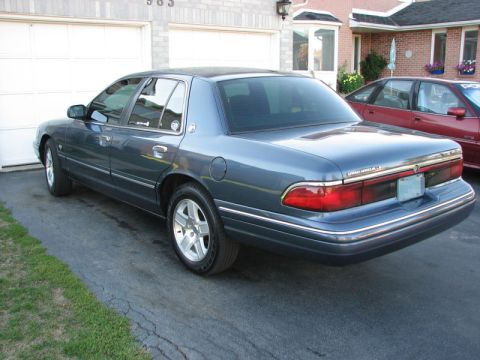 1985 Mercury Grand Marquis - This blue sedan is seen from the rear 3/4 angle, with another red sedan visible in the background.