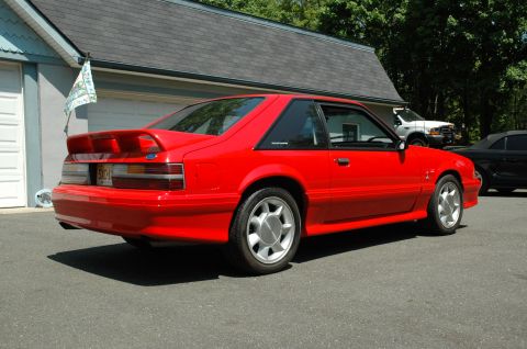 1993 Ford Mustang - A red 1993 Ford Mustang is parked in a driveway next to a garage, with other vehicles visible in the background.