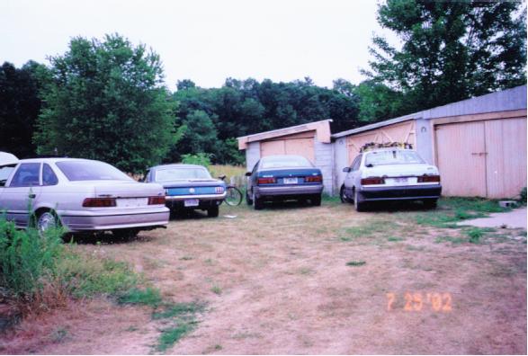 Four cars including a white sedan, white Mustang, and green sedan parked on grass by wooden garages, dated 7-25-02