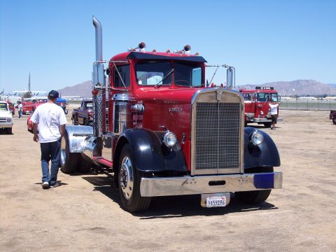 1981 Kenworth W900A - A classic red and black semi-truck is parked outdoors, with other vehicles and a person walking nearby under a clear blue sky.
