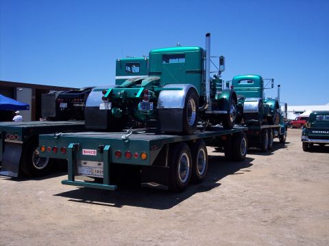 1981 Kenworth W900A - Multiple semi-truck cabs are being transported on a flatbed trailer under a clear blue sky.