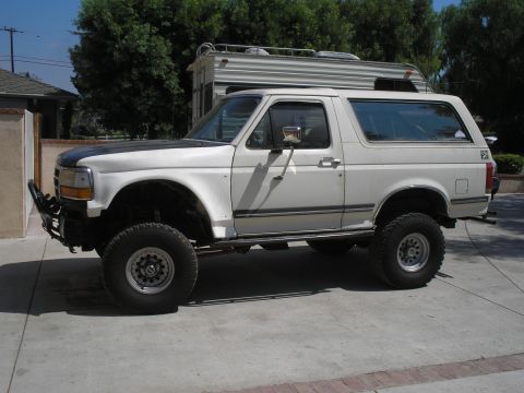 1990 Ford Bronco - Driver's-side profile of a white lifted Ford Bronco with hardtop parked on a residential concrete driveway, a travel trailer behind it. It…