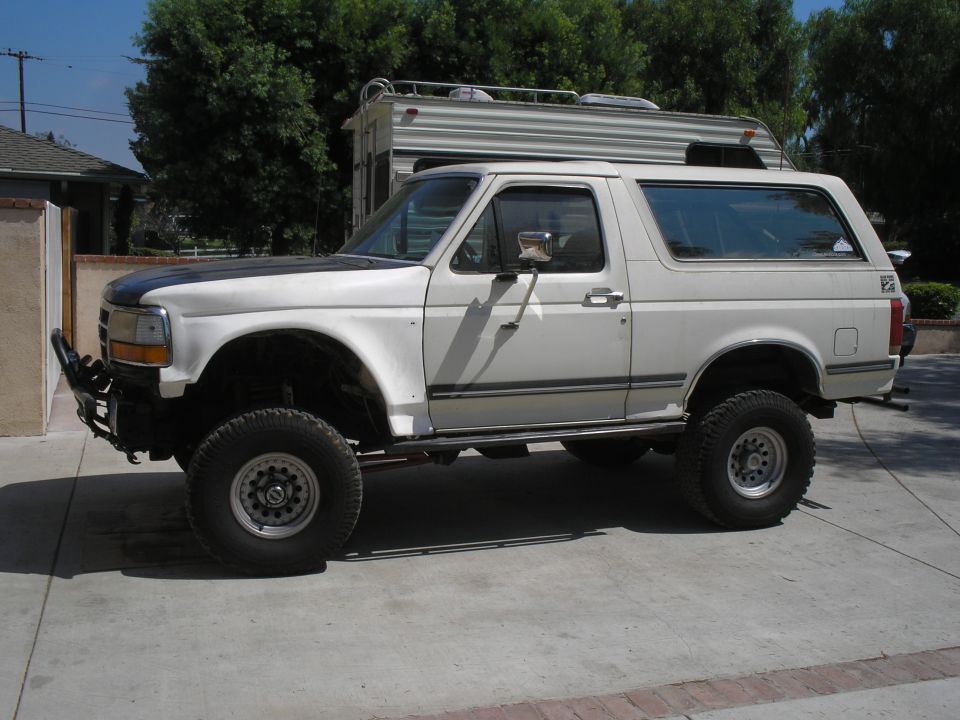 Driver-side profile of a white lifted Ford Bronco on a driveway with a travel trailer behind