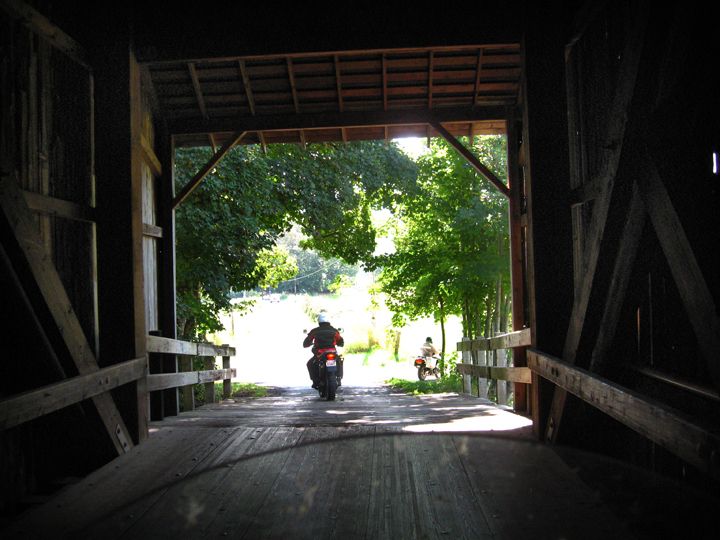 Two motorcycles ride out of a wooden covered bridge into a sunny, tree-lined road