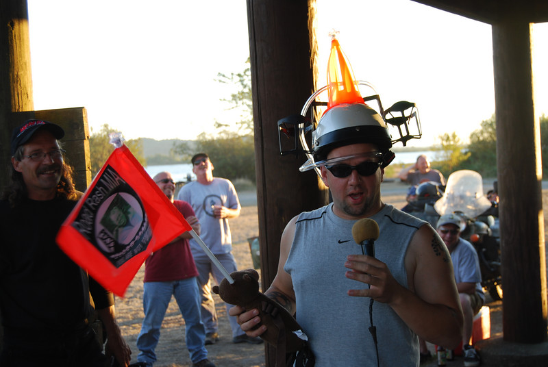 Man with novelty helmet holding microphone and toy, people in background, red flag with text