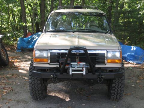 1979 Ford Bronco - This tan SUV features a prominent winch and bull bar on its front end, set against a wooded background.