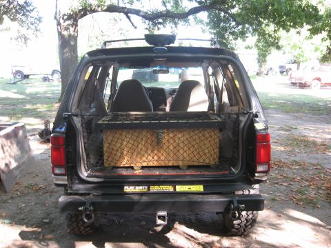 1979 Ford Bronco - Rear view of a black SUV with its cargo area open, revealing a large box covered in netting.