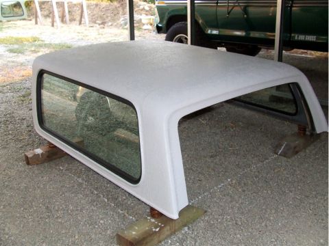1979 Ford Bronco - A white camper shell sits on wooden blocks, with a green pickup truck visible in the background.