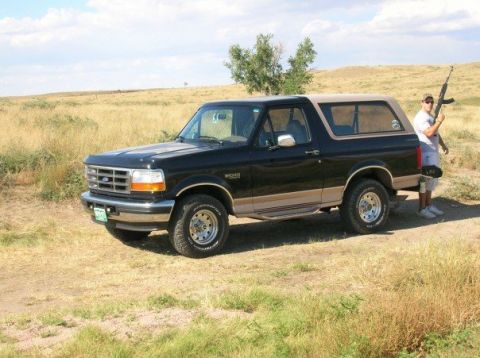 1996 Ford Bronco - A person stands with a rifle next to a black Ford Bronco in a grassy, open field under a partly cloudy sky.