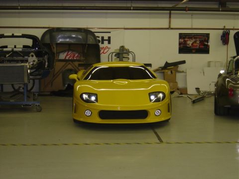 1996 Ford Bronco - A yellow sports car is parked in a workshop with other vehicles and parts around.