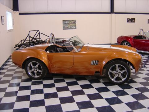 1996 Ford Bronco - Side view of an orange AC Cobra replica in a garage with a checkered floor and another car in the background.