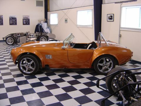 1996 Ford Bronco - An orange convertible sports car with a white stripe is parked indoors next to a chassis.