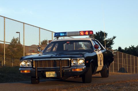 1973 Dodge 1500 - A police car with a light bar is parked on a dirt road near a chain-link fence.
