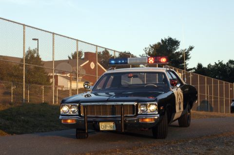 1973 Dodge 1500 - This police car, likely from the 1970s, is parked outdoors near a chain-link fence and a building.