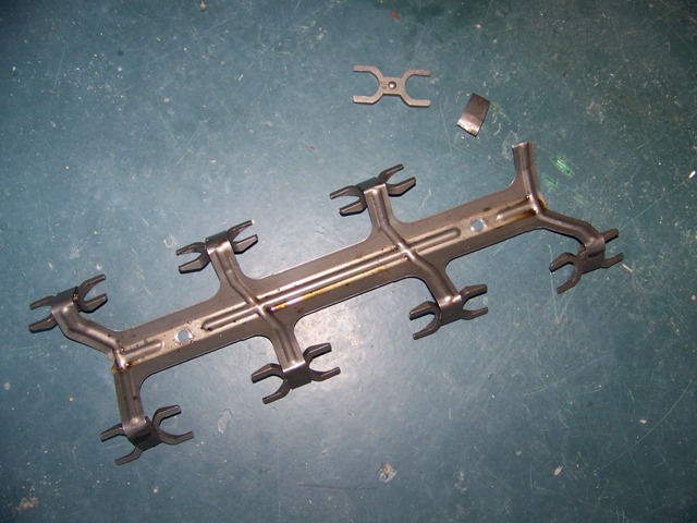 Stamped steel rocker arm retainer rail with forked ends and a broken-off piece on a green floor