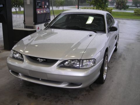 1998 Ford Mustang - A silver 1998 Ford Mustang coupe is parked at a gas station, with raindrops visible on the car's surface.