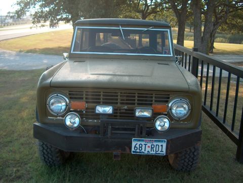 1979 Ford Bronco - This classic Ford Bronco is parked on grass next to a black fence, with trees and a road in the background.