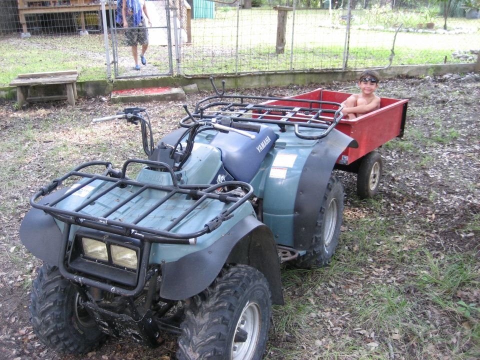 Teal Yamaha ATV with cargo racks towing a red utility trailer with a child inside, in a fenced yard