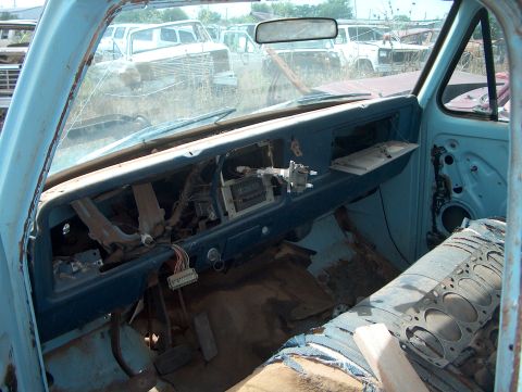 1979 Ford Bronco - View from inside a dilapidated truck cab, showing a damaged dashboard and worn seats amidst a junkyard setting.