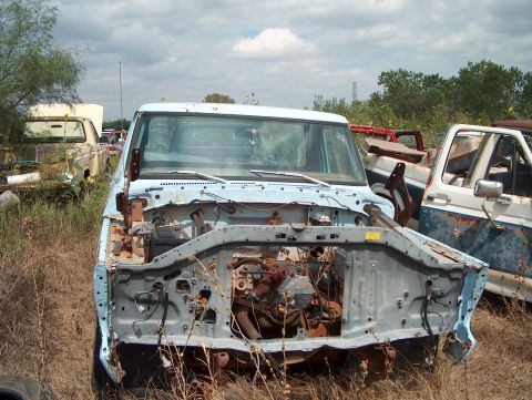 1979 Ford Bronco - A light blue truck sits in a junkyard, its front end dismantled, revealing its engine and chassis.