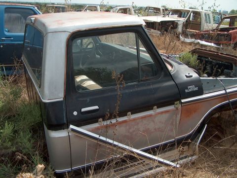 1979 Ford Bronco - Junkyard scene the side of a pickup truck cab and a camper shell, surrounded by other abandoned vehicles.