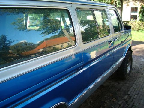 1978 Ford E250 - A blue and silver van is seen from the side, with a reflection of a white van in its window.