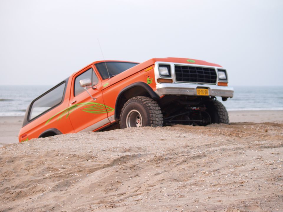 Orange 1979 Ford Bronco stuck on a sandy beach with the ocean in the background