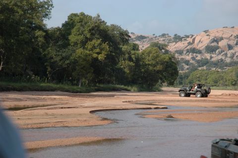 2001 Jeep Wrangler - A Jeep Wrangler navigates a dry riverbed, surrounded by trees and distant rocky terrain.