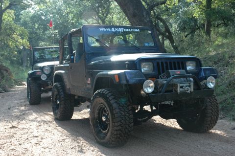 2001 Jeep Wrangler - Two dark blue Jeep Wranglers with off-road modifications drive down a dirt path surrounded by trees.