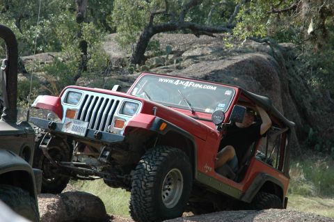 2001 Jeep Wrangler - A red Jeep Wrangler is shown off-roading on a rocky trail, with another vehicle partially visible to the left.