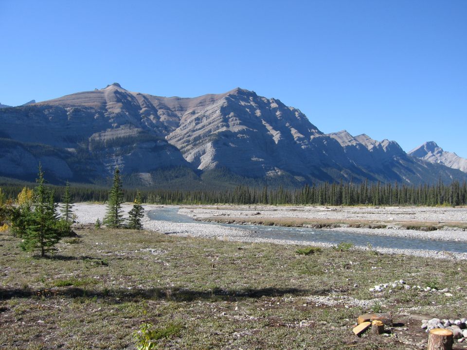 Mountain range with layered rock formations, a riverbed with a stream, and scattered pine trees under a clear blue sky