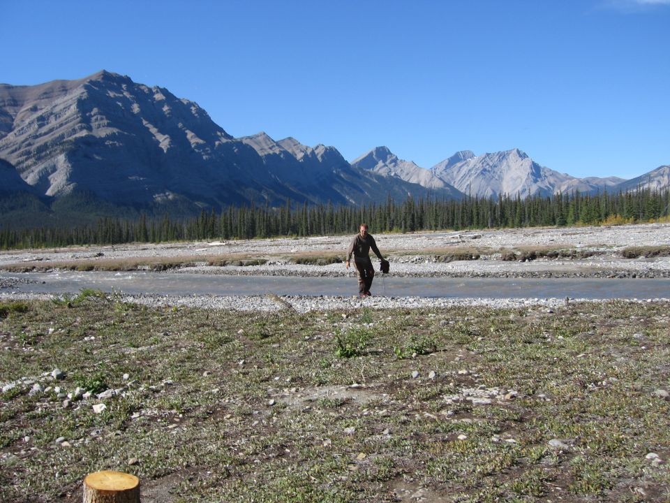 Person walking across a rocky riverbed with mountains and pine trees in the background
