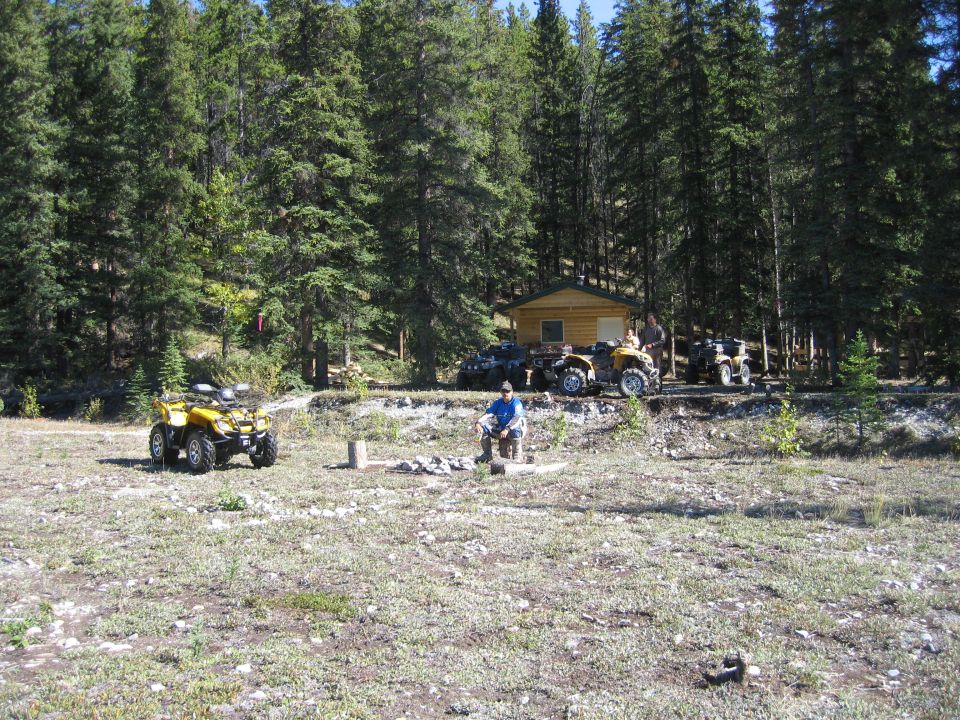 Several ATVs are parked in a clearing in front of a small wooden cabin, surrounded by pine trees. A person sits on a rock in the foreground