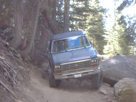 1986 Ford Bronco - A dark blue SUV navigates a rocky, wooded trail, with another vehicle partially visible behind it.