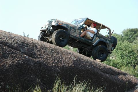 2001 Jeep Wrangler - A man drives a camouflage Jeep Wrangler up a steep, rocky incline on a sunny day.