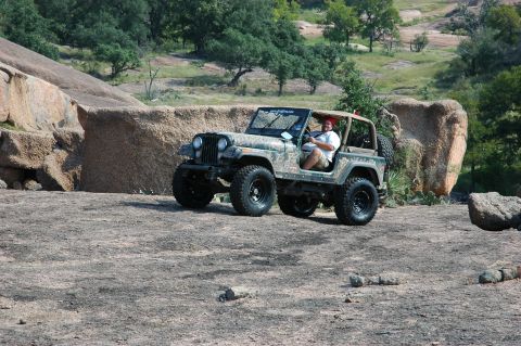 2001 Jeep Wrangler - A man drives a camouflaged Jeep CJ-7 through a rocky, outdoor landscape.