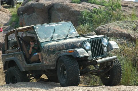 2001 Jeep Wrangler - A Jeep CJ-7 with camouflage paint navigates a rocky terrain, with a website visible on its windshield.