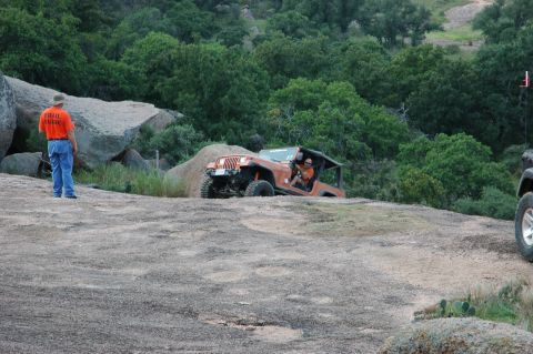 2001 Jeep Wrangler - A man in an orange shirt with 'TRAIL GUIDE' text stands near an orange Jeep Wrangler driving on a rocky hill.