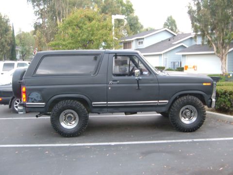 1981 Ford Bronco - Side view of a dark gray Ford Bronco with off-road tires, parked in a lot with buildings in the background.
