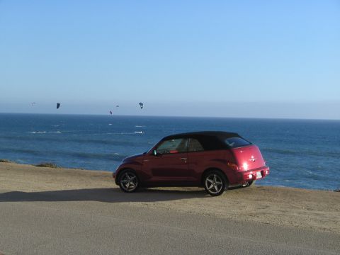 2005 Chrysler PT Cruiser - A red convertible PT Cruiser is parked by the ocean, with kitesurfers visible on the water.