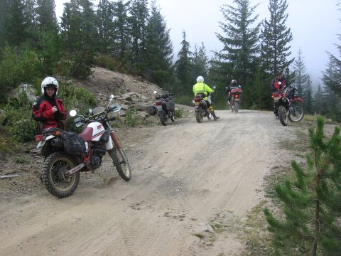 1979 Ford Bronco - Riders on motorcycles navigate a dirt road through a misty, wooded mountain landscape.