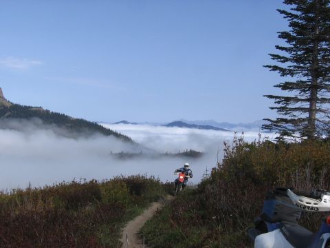 1979 Ford Bronco - A lone motorcyclist navigates a winding dirt path through a mountainous landscape shrouded in fog.