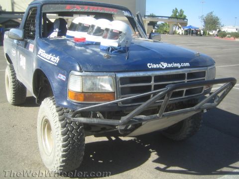 1990 Ford Bronco - Dark blue Ford off-road race truck numbered 340 with crash helmets on the cowl and a steel tube front bumper, parked on pavement.