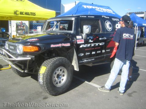 1990 Ford Bronco - Black Toyota FJ Cruiser off-road race truck with Nitto and KC graphics parked under event canopies as a man in an ORW shirt stands nearby.