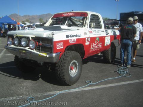 1990 Ford Bronco - Red and white Chevrolet off-road race truck #304 with KC lights and BFGoodrich graphics parked in a desert paddock with crew nearby.